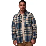 A man wearing a plaid button-up shirt with blue, cream, and navy checkered patterns over a light beige shirt and tan pants.