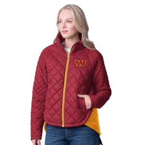 A woman wears a quilted NFL jacket in burgundy and gold. It features a \“W\“ logo on the chest, yellow zipper, curved hem, and a contrasting yellow inner lining, paired with jeans.