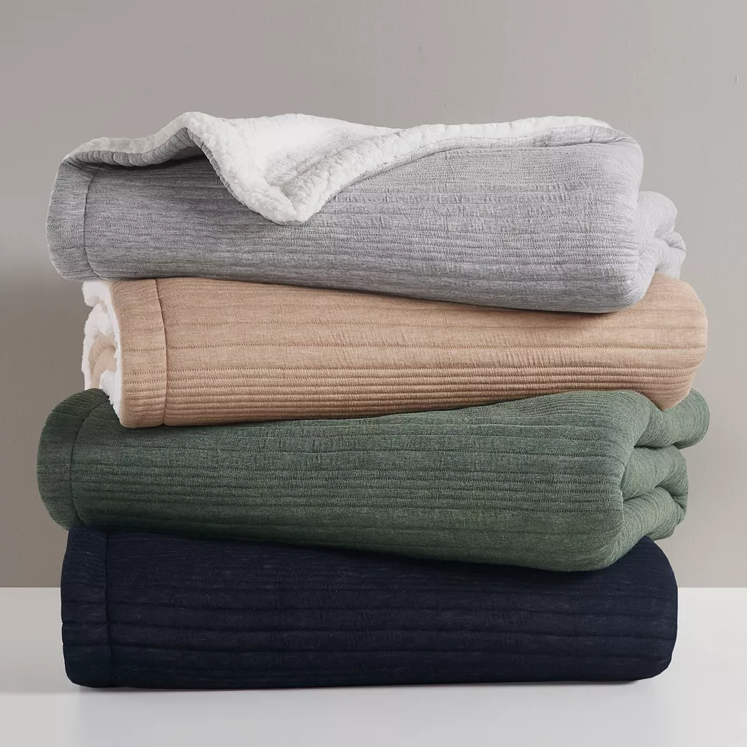 A stack of four textured blankets in white, beige, green, and blue, with the top one displaying a white sherpa lining.