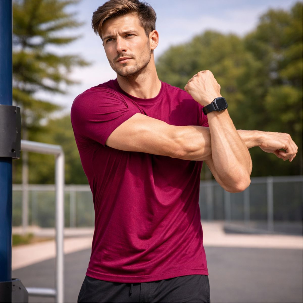 A man in a maroon workout shirt stretches his arm while wearing a black smartwatch, standing in an outdoor fitness park.