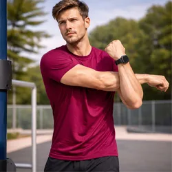 A man in a maroon workout shirt stretches his arm while wearing a black smartwatch, standing in an outdoor fitness park.