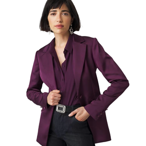 A woman is wearing a WHBM Fully-Lined Blazer in a deep purple color, paired with a matching shirt and dark jeans. The blazer is styled with a decorative belt and statement earrings.