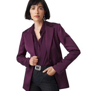 A woman is wearing a WHBM Fully-Lined Blazer in a deep purple color, paired with a matching shirt and dark jeans. The blazer is styled with a decorative belt and statement earrings.