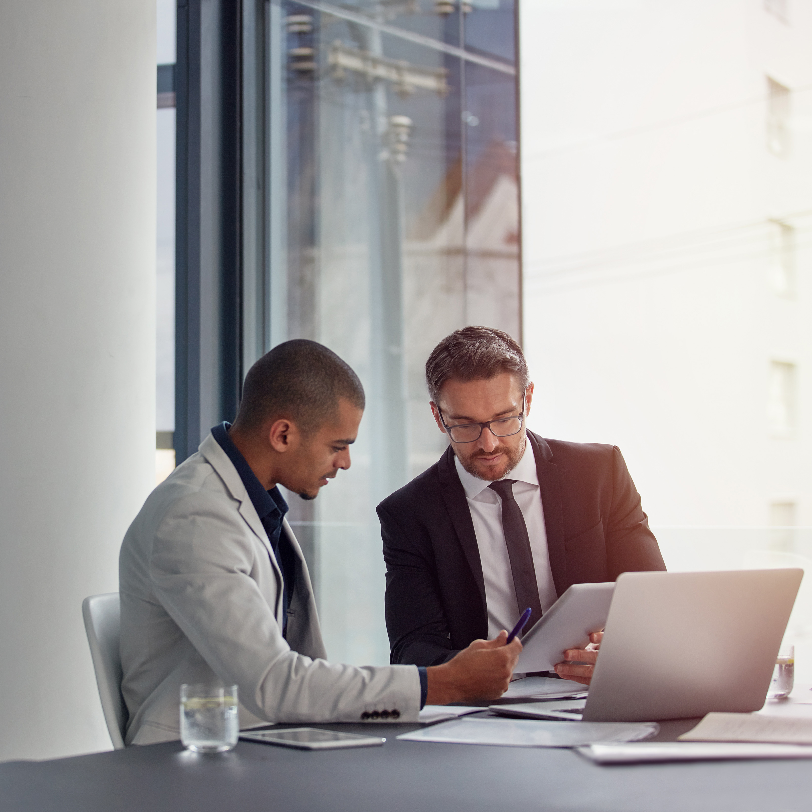 Two men reviewing a document on a digital tablet with a laptop open on the desk.