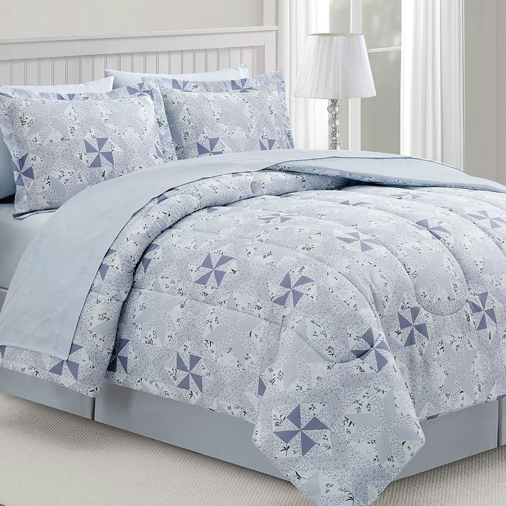 A bed is covered with a comforter and pillowcases featuring a light blue and white geometric pattern with star-like designs. A lamp with a white shade is on a nearby nightstand.