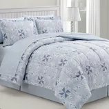 A bed is covered with a comforter and pillowcases featuring a light blue and white geometric pattern with star-like designs. A lamp with a white shade is on a nearby nightstand.