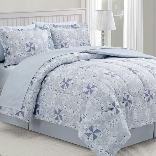 A bed is covered with a comforter and pillowcases featuring a light blue and white geometric pattern with star-like designs. A lamp with a white shade is on a nearby nightstand.