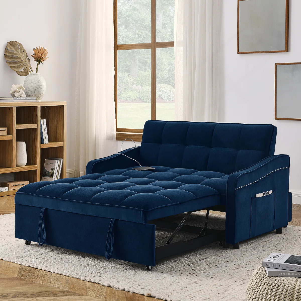 Blue upholstered sofa bed with a pull-out sleeper design. Features a side pocket and nailhead trim, positioned in a cozy living room setup with a window, rug, and bookshelf.