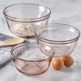 Three transparent mixing bowls, one with flour, are placed alongside a wooden spoon, a towel, and three eggs on a marble surface.