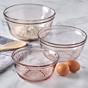 Three transparent mixing bowls, one with flour, are placed alongside a wooden spoon, a towel, and three eggs on a marble surface.