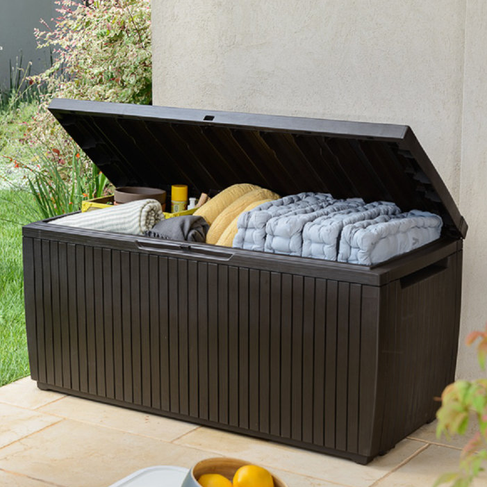 80-Gallon Deck Box Bench with a hinged lid, ideal for storing outdoor items like cushions and blankets. Features a slatted design and is positioned on a patio next to a garden.