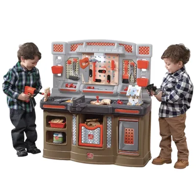 Children are playing with a toy workbench set featuring tools like a drill, saw, pliers, and a vise, along with a small wooden house project.