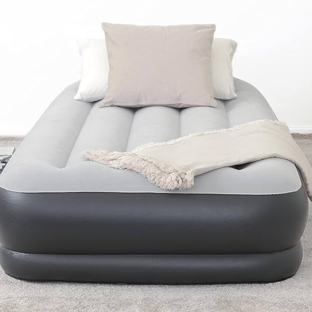 Gray and black inflatable air mattress with a soft top surface, featuring two pillows and a fringed throw blanket, placed on a carpeted floor.