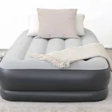 Gray and black inflatable air mattress with a soft top surface, featuring two pillows and a fringed throw blanket, placed on a carpeted floor.