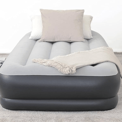 Gray and black inflatable air mattress with a soft top surface, featuring two pillows and a fringed throw blanket, placed on a carpeted floor.