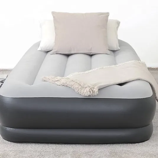 Gray and black inflatable air mattress with a soft top surface, featuring two pillows and a fringed throw blanket, placed on a carpeted floor.
