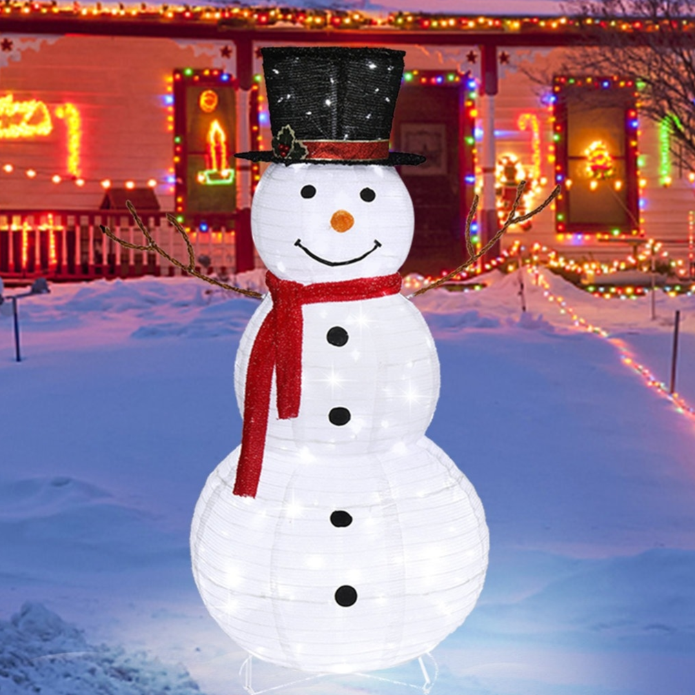 LED snowman decoration with a top hat and red scarf, in a snowy yard with colorful Christmas lights adorning a house in the background.
