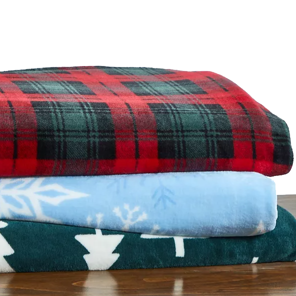 A stack of three holiday-themed velvety throws, featuring a red and black plaid pattern on top, a light blue one with white snowflakes in the middle, and a dark green one with white tree silhouettes at the bottom.