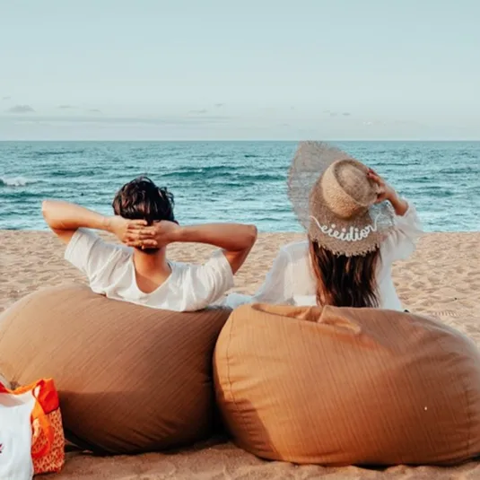 Two people sit on large, brown bean bags facing the ocean on a beach. One wears a wide-brimmed straw hat.