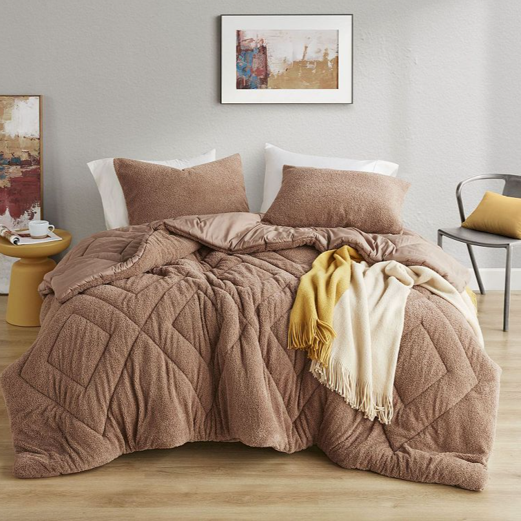 Cozy bedroom setup with a textured brown comforter, matching pillows, a contrasting yellow and white throw, and abstract artwork. Includes a side table with a decorative piece and a modern chair with a yellow cushion.
