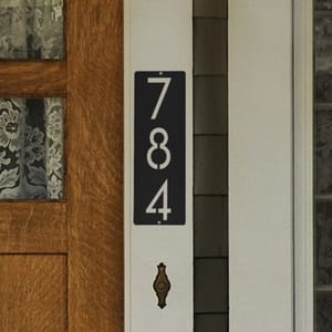 Vertical house number sign with the digits 7, 8, and 4 displayed on a black background, mounted beside a wooden door with lace curtains.