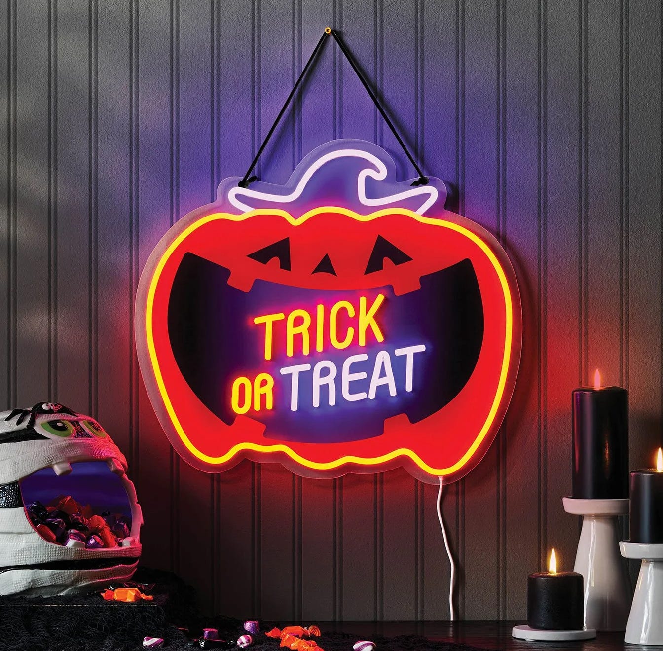 Neon pumpkin sign with \“Trick or Treat\“ message, accompanied by black candles and a mummy-themed candy holder filled with sweets.