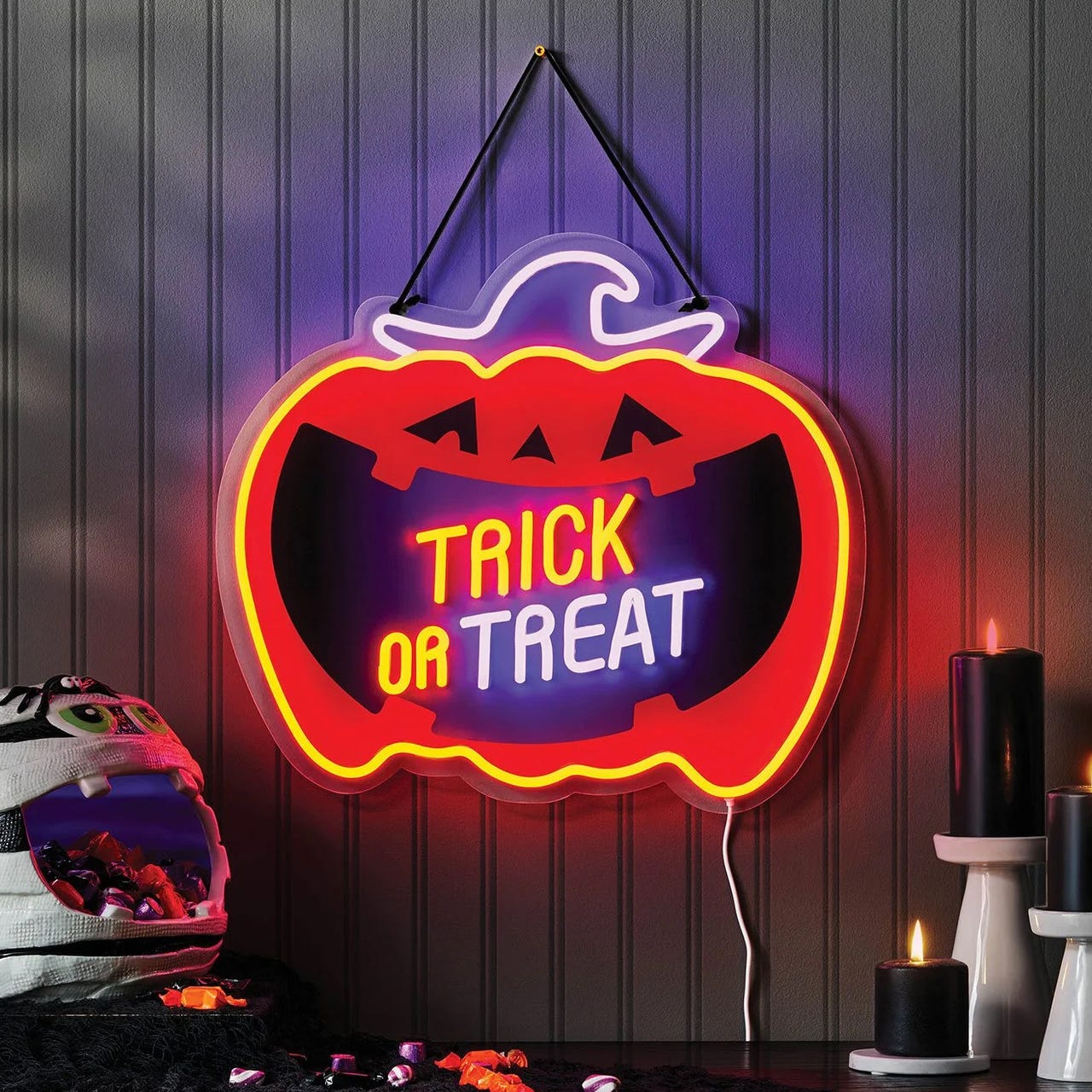 Neon pumpkin sign with \“Trick or Treat\“ message, accompanied by black candles and a mummy-themed candy holder filled with sweets.