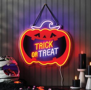 Neon pumpkin sign with \“Trick or Treat\“ message, accompanied by black candles and a mummy-themed candy holder filled with sweets.