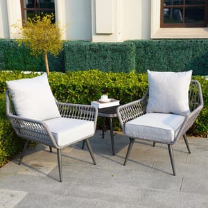 Outdoor seating set with two gray wicker chairs featuring white cushions and a small side table in between, set on a patio with greenery in the background.