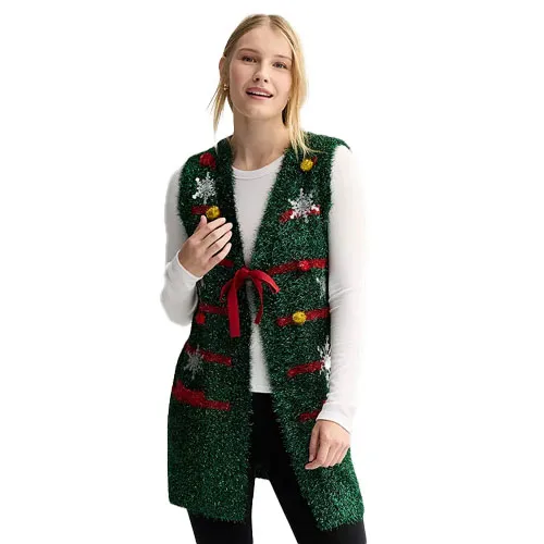A woman is wearing a green, fuzzy Christmas-themed vest with decorative elements and a red tie, layered over a white long-sleeve shirt and black pants.