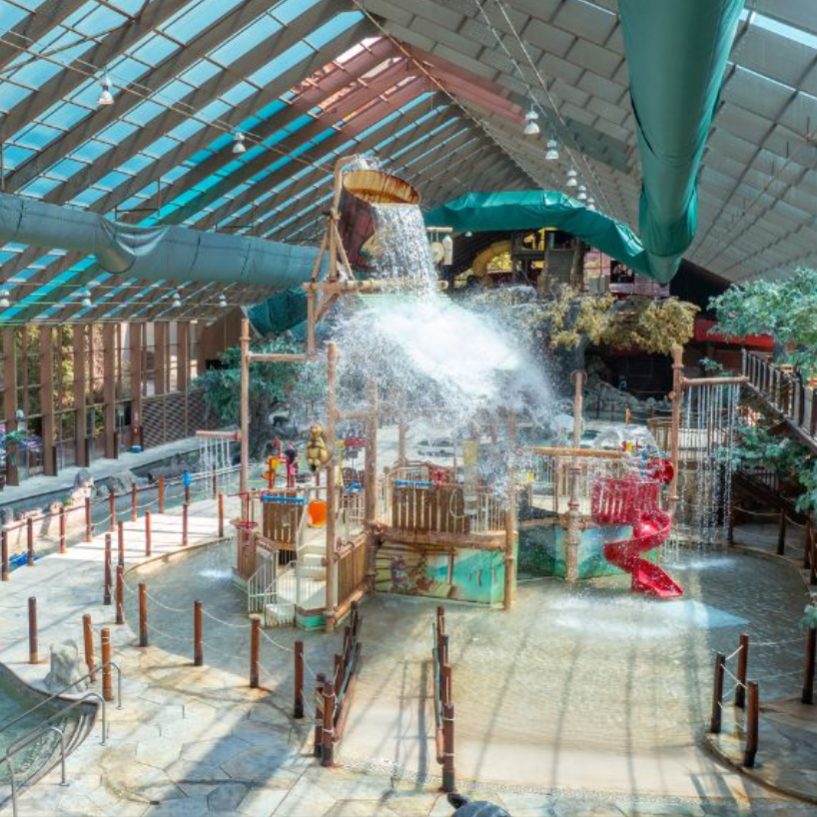 An indoor water park featuring a splash play area with water features, a large tipping bucket, a red spiral slide, and a lazy river with rocks and animal sculptures under a glass ceiling.