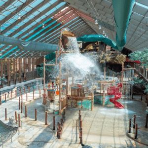 An indoor water park featuring a splash play area with water features, a large tipping bucket, a red spiral slide, and a lazy river with rocks and animal sculptures under a glass ceiling.