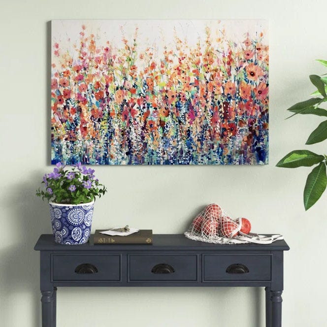 A vibrant painting featuring a dense field of colorful flowers with abstract brushstrokes. It hangs above a dark blue console table, which holds a potted plant, a book, and netted decor.