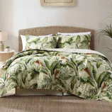 A bedding set with a banana leaf print, featuring a woven headboard, a side table with a lamp, and a small plant.