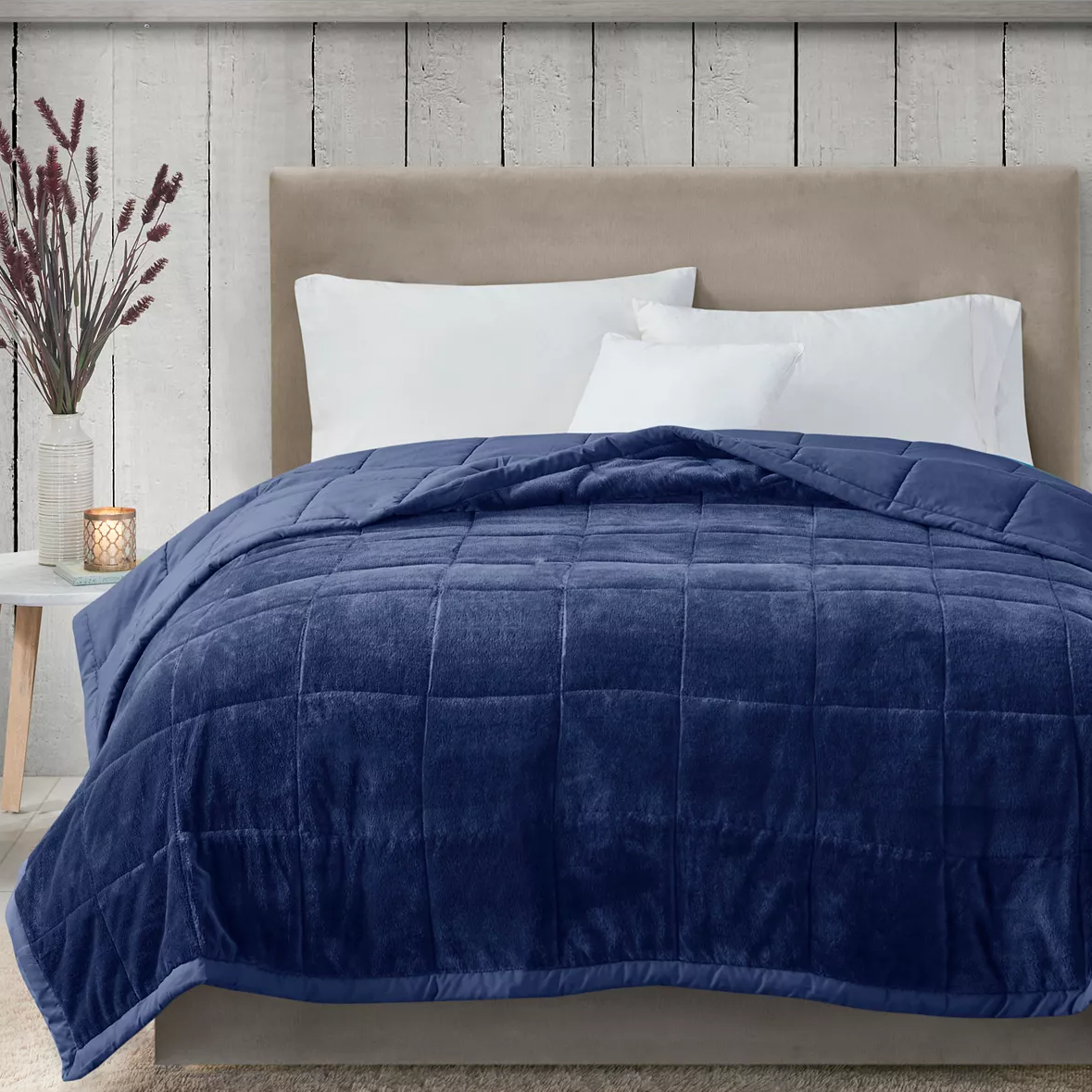 A blue comforter on a bed, with a pair of white pillows and a small bedside table with a candle and vase.