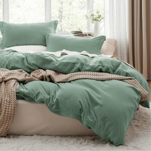 A neatly made bed features green pillowcases and duvet cover, accompanied by a beige textured blanket. Bright natural light filters through large windows, illuminating a serene bedroom setting with a soft rug and flowers on a side table.