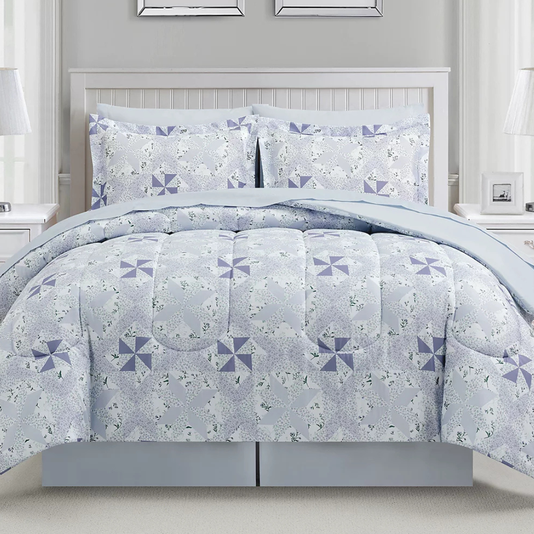 A bed is dressed with a light blue and lavender quilt featuring a pinwheel pattern, accompanied by matching pillow shams. The room includes white side tables and a white paneled headboard.