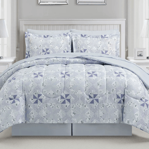 A bed is dressed with a light blue and lavender quilt featuring a pinwheel pattern, accompanied by matching pillow shams. The room includes white side tables and a white paneled headboard.