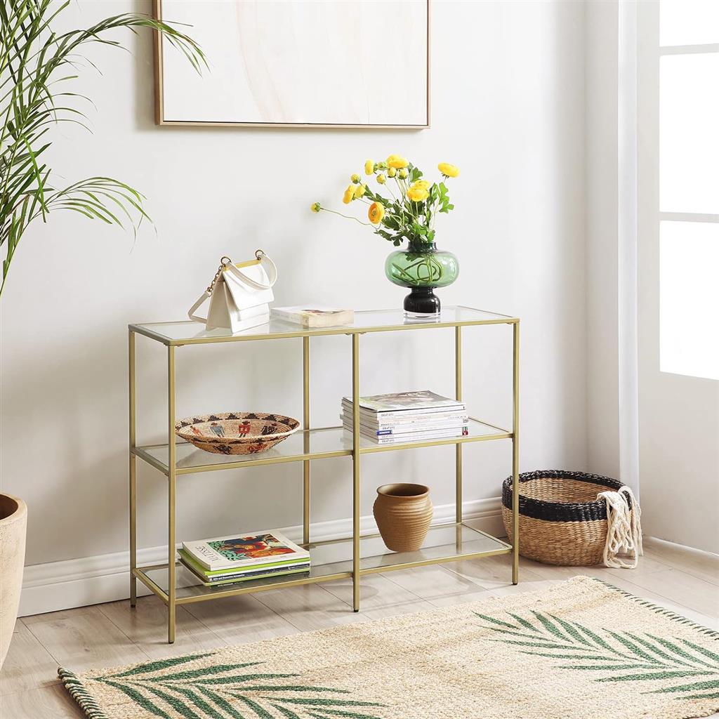 Gold metal console table with glass shelves displaying books, a green vase with flowers, a bowl, and a small white purse.