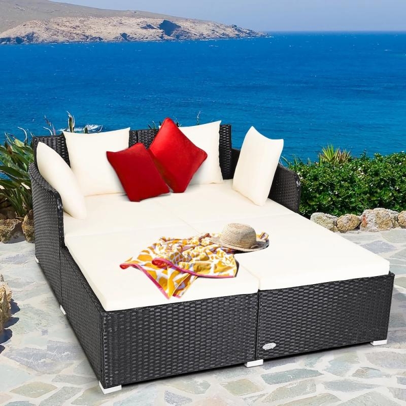 Wicker patio daybed with white cushions and contrasting red pillows, set by a seaside, includes a hat and patterned cloth accessory.