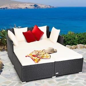 Wicker patio daybed with white cushions and contrasting red pillows, set by a seaside, includes a hat and patterned cloth accessory.