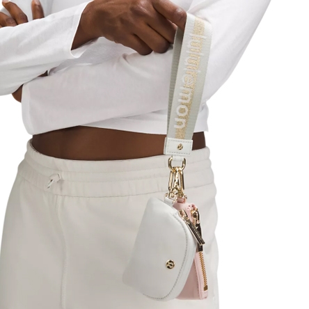 A small white and pink pouch with a gold zipper, attached to a strap with \“lululemon\“ branding, is worn on the wrist.