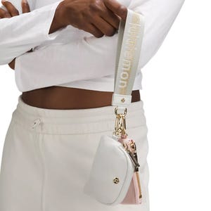 A small white and pink pouch with a gold zipper, attached to a strap with \“lululemon\“ branding, is worn on the wrist.