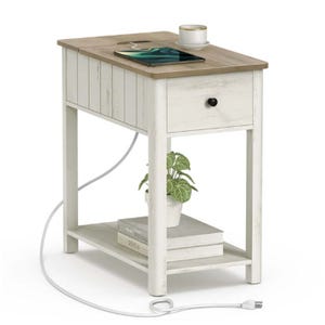 A small white wooden side table with a light brown top featuring a drawer, an open shelf holding books and a small plant, and a built-in charging outlet with a cord. A cup and a tablet are placed on top.