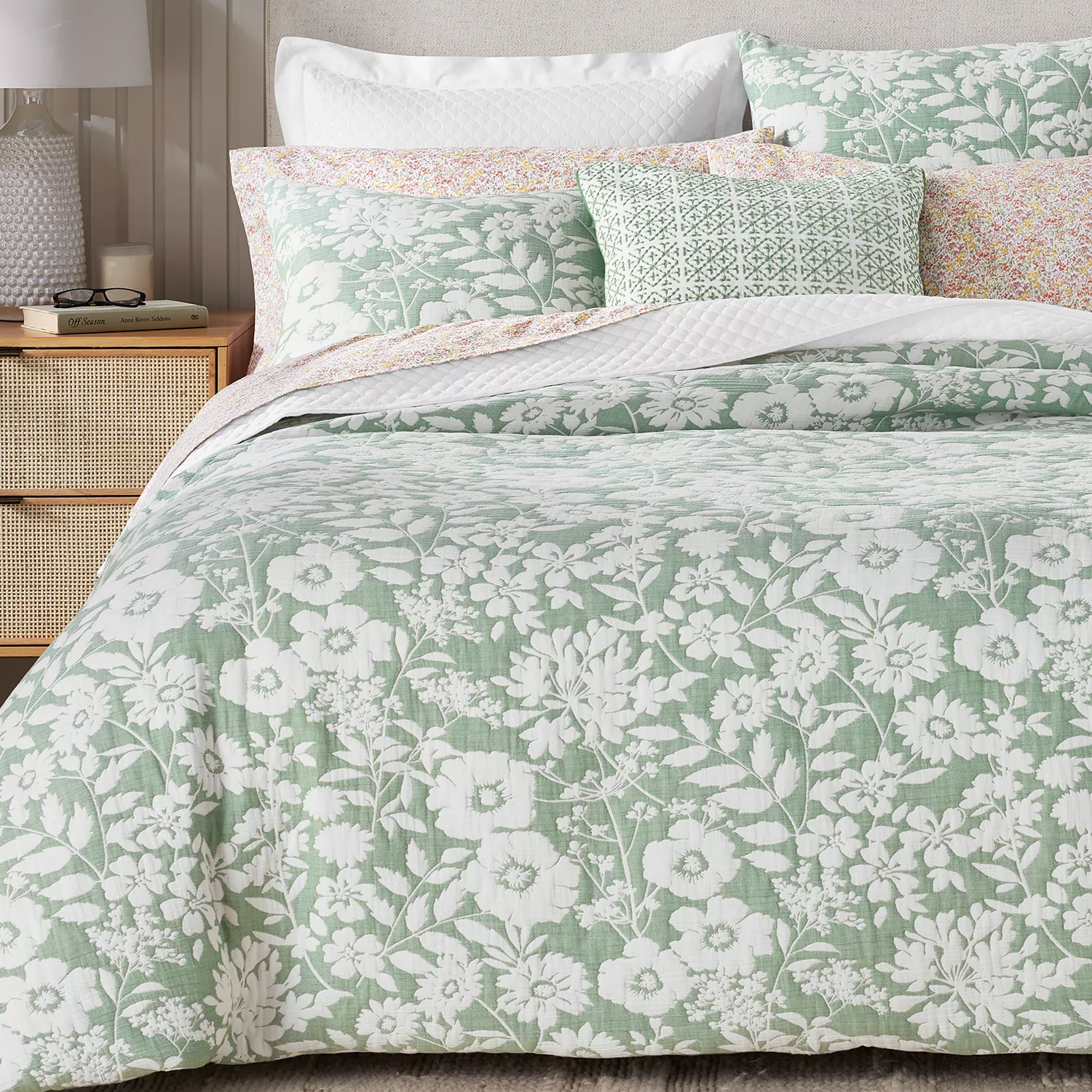 A bed is shown with a quilt set featuring a green and white floral design, complemented by pillows in similar patterns and pastel colors. A bedside table with a lamp and a book is visible.