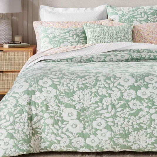 A bed is shown with a quilt set featuring a green and white floral design, complemented by pillows in similar patterns and pastel colors. A bedside table with a lamp and a book is visible.