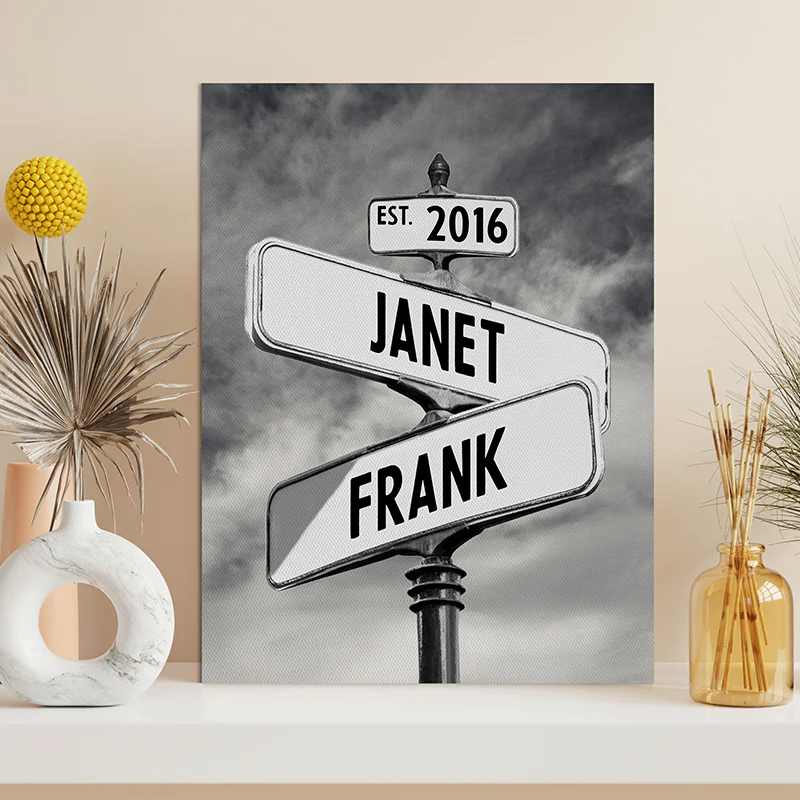 A black and white canvas print features a street sign design with the names \“Janet\“ and \“Frank\“ intersecting, accompanied by the text \“EST. 2016,\“ placed on a shelf with decorative vases and dried plants.