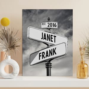 A black and white canvas print features a street sign design with the names \“Janet\“ and \“Frank\“ intersecting, accompanied by the text \“EST. 2016,\“ placed on a shelf with decorative vases and dried plants.