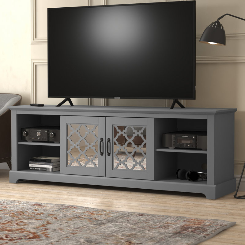 A modern gray TV stand with decorative cabinet doors, housing electronic devices and books, with a flat-screen TV on top and a floor lamp beside it.