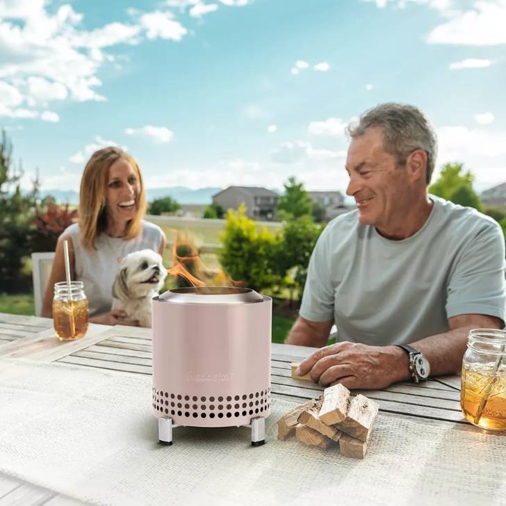 A portable outdoor fire pit and a pair of wooden logs on a patio table with two smiling people and a dog in the background.